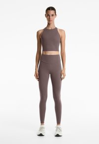 Top corto marrone con scollo a halter e leggings high-waisted in un tessuto morbido e elasticizzato. Il modello indossa scarpe da ginnastica bianche, in piedi contro uno sfondo bianco.