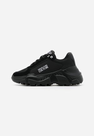 Sort chunky sneaker med tyk sål, snørebånd foran og "Versace Jeans Couture" logo på siden og tungen, set fra siden på hvid baggrund.