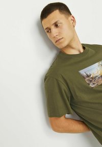 Olivengrøn bomulds T-shirt med korte ærmer, der har et rektangulært grafisk print af et landskab på forsiden.