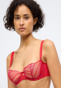 Sutiã vermelho de rendinha com copas em malha transparente, suporte de aros e alças ajustáveis. Apresenta padrões bordados intricados ao longo das bordas.