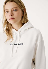 Jonge vrouw met blond haar draagt een witte hoodie met "RED BULL RACING" geborduurd op de borst tegen een vlakke lichte achtergrond.