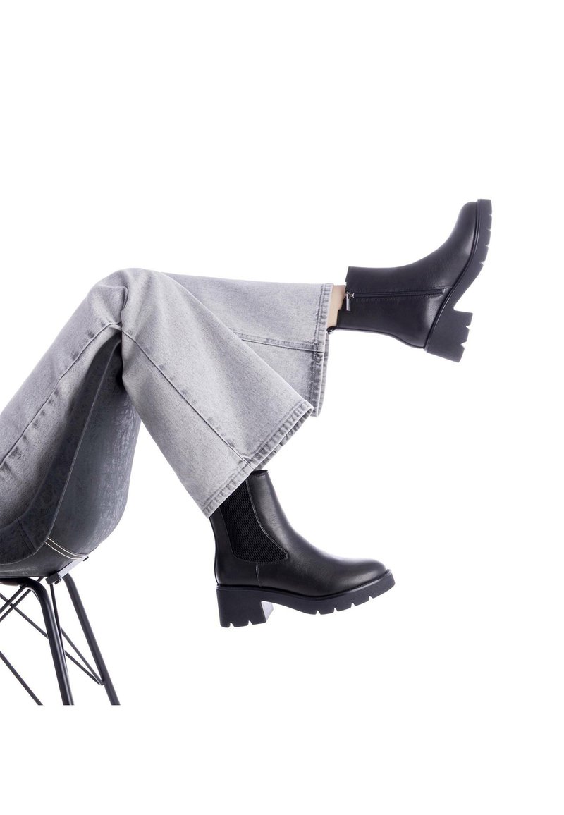 Botas negras de tobillo con un panel texturizado, tacón grueso y un cierre lateral, combinadas con unos jeans de pierna amplia en gris claro. El entorno es minimalista.