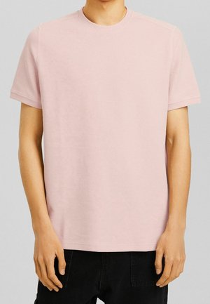 Mann trägt ein hellrosa kurzärmeliges Rundhalsshirt und schwarze Hose, steht vor einem schlichten grauen Hintergrund.