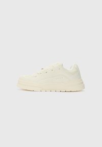 White sneaker with textured upper, rounded toe, flat laces, and thick rubber sole. Subtle stitching and ventilation holes on the sides.