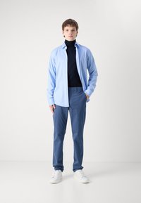 Lyseblå langermet skjorte over en svart genser med høyt liv, kombinert med blå bukser og hvite joggesko. Glatt stoff, avslappet passform og enkelt design.
