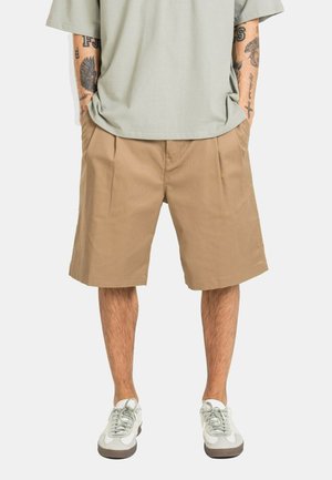Man wearing beige knee-length shorts, light gray T-shirt, and white sneakers, standing with hands in pockets against plain background.