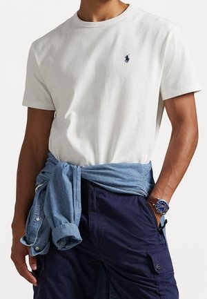 Mand iført hvid kortærmet T-shirt med lille broderet logo, blå cargobukser, denimskjorte bundet om livet og armbåndsur.