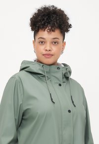 Πράσινο αδιάβροχο μπουφάν με φερμουάρ μπροστά, κουμπιά, ρυθμιζόμενη κουκούλα με κορδόνι και λεία υφή. Διαθέτει μαύρες μεταλλικές λεπτομέρειες.