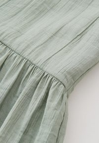 Gros plan sur un tissu vert clair rassemblé au niveau d'une couture, montrant une fine texture verticale et un matériau doux et fluide.
