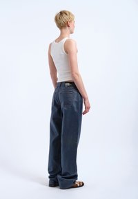 Breda mörkblå jeans med en urtvättad textur, sedda bakifrån. En vit åtsittande tanktopp och mönstrade skor fulländar looken.