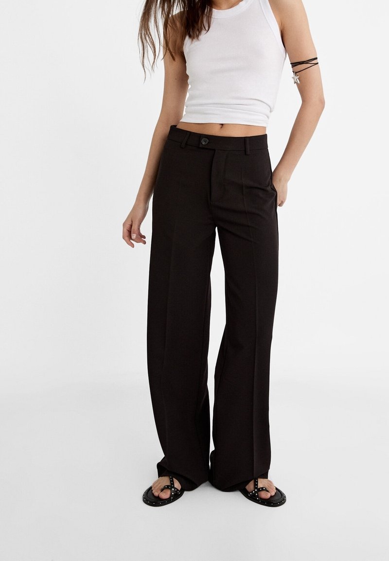 Pantalon noir à jambe large avec fermeture à bouton, passants de ceinture et texture lisse. Associé à un débardeur blanc ajusté et des sandales simples.