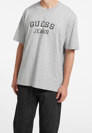 Szara bawełniana koszulka z krótkim rękawem z napisem "GUESS JEANS" w czerni, o luźnym kroju, zestawiona z ciemnymi dżinsami.