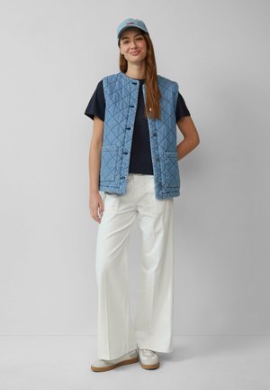Gequilted blauwe denim vest met een ronde halslijn, voor knopen en twee zakken, gedragen over een zwart shirt en gecombineerd met witte wijde pantalon.