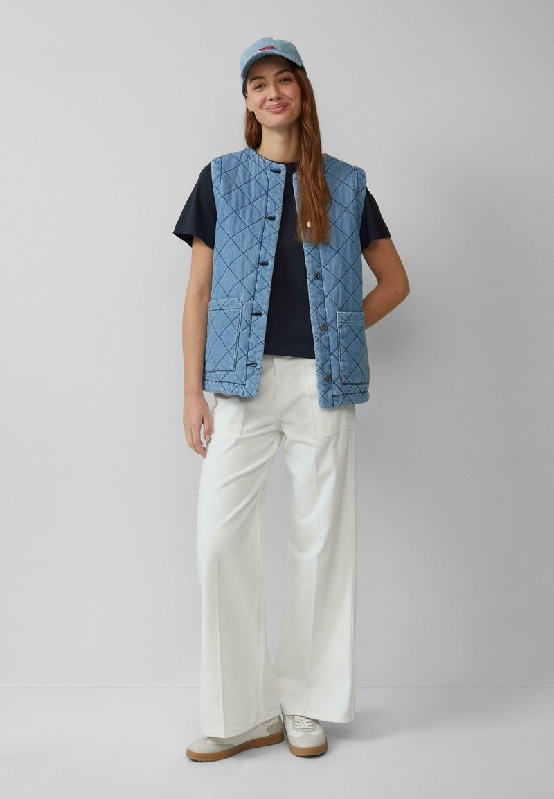 QD puchowa niebieska kamizelka dżinsowa z okrągłym dekoltem, z przodu z guzikami i dwoma kieszeniami, noszona na czarnej koszuli, zestawiona z białymi spodniami o szerokich nogawkach.