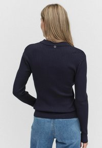 Marineblauwe geribde trui met lange mouwen en een kraaghals. Heeft een subtiele cirkelvormige accenten aan de achterkant. Materiaal lijkt zacht en gestructureerd.