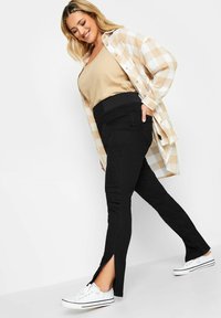 Svarta slim fit byxor med sidofickor, kombinerat med en beige linne och en ljus rutig skjorta. Vita sneakers fulländar outfiten.