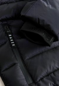 Sort pufferjakke tæt på med en sidezipper og en tekstureret ribbet manchet med præget branding. Glat, skinnende materiale med kviltede paneler.