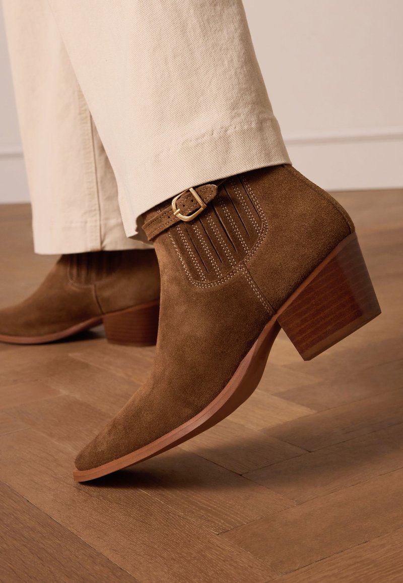 Stivale ankle in suede marrone con punta affusolata, cuciture decorative e un accento in metallo dorato, dotato di tacco in legno impilato.