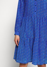 Blå klänning med blommönster, långa ärmar, knappar fram, och lager av kjolens nederkant. Tillverkad av lättviktig tyg.