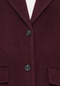 Burgundia villasegu segu blazer, mille on nurgeline krae, kaks esipocketit ja mustad nupud. Kangas on pehme tekstuuriga ja veidi karvane välimus.