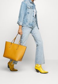 Persona con giacca di denim chiaro e jeans che cammina, portando una grande borsa tote gialla senape, indossando stivaletti gialli brillanti con tacco.
