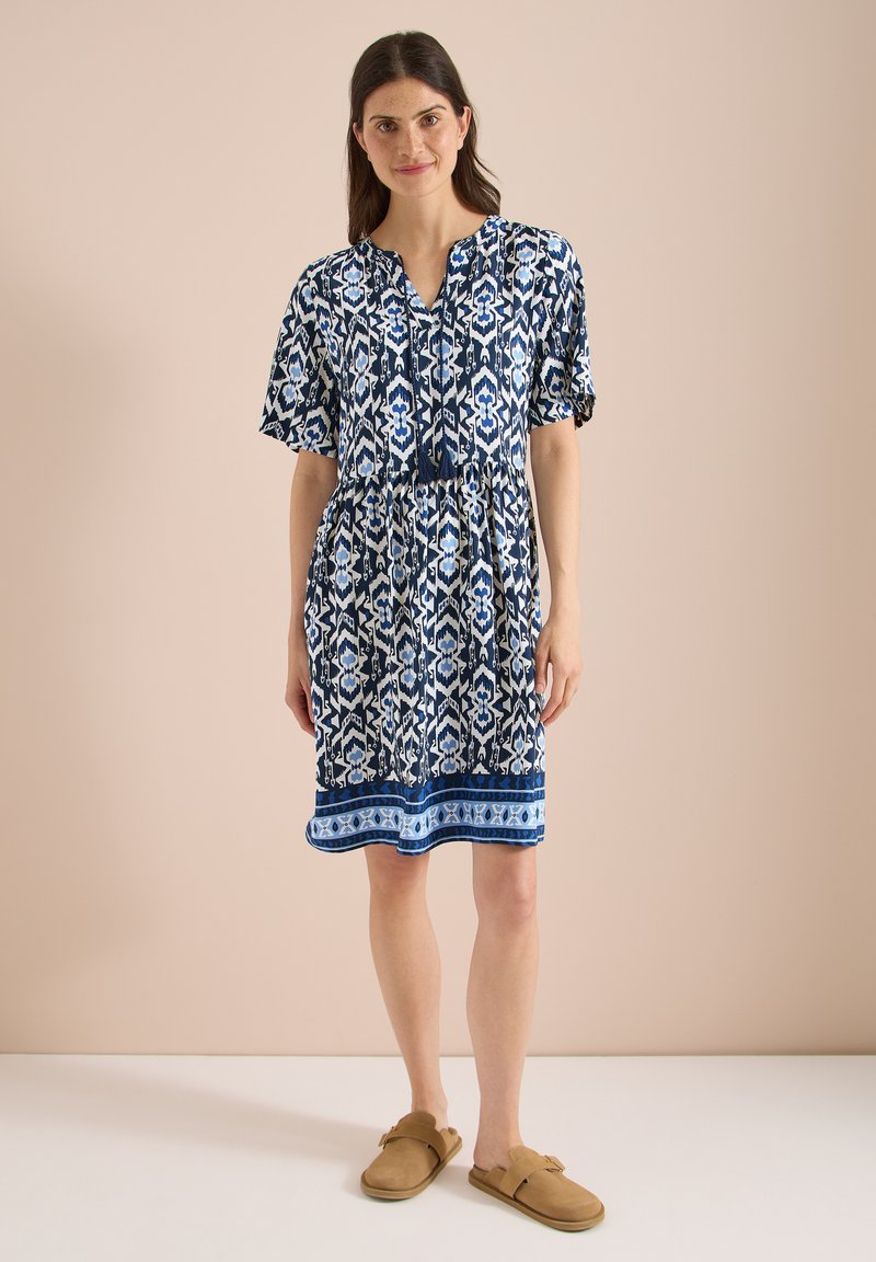 Femme aux cheveux foncés portant une robe à motifs bleus, blancs et noirs longueur genou et des chaussures beige à enfiler, debout sur un fond uni.