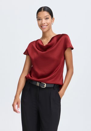 Jeune femme souriante portant un chemisier en satin rouge et un pantalon noir avec une ceinture noire, debout avec une main dans la poche sur fond blanc.
