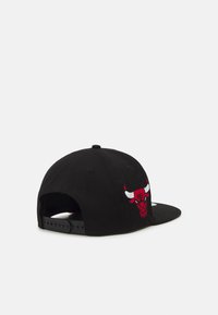 Gorra negra con visera plana, con un logo de toro bordado en rojo en el panel frontal izquierdo. Correa de plástico ajustable en la parte trasera.