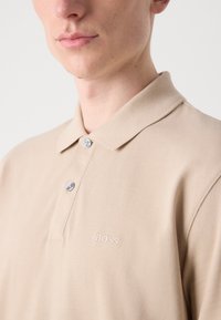 Bežinė polo marškinėliai, pagaminti iš tekstūruotos medvilnės. Yra klasikinis apykaklė, trys sagos ir siuvinėtas "BOSS" logotipas krūtinėje.