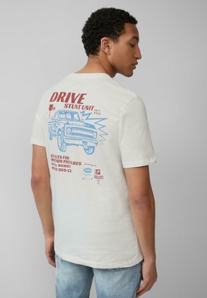 Witte katoenen t-shirt met rode en blauwe tekst en een afbeelding van een vintage vrachtwagen op de achterkant, met extra sponsorinformatie en accenten.