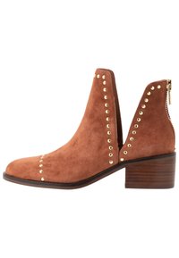 Bottines en daim marron avec un bout rond, découpes latérales et accents en clous dorés le long des bords. Présente un talon en bois et une fermeture éclair.