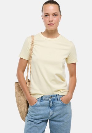 Jeune femme portant une chemise jaune pâle à manches courtes et un jean bleu, les mains dans les poches, avec un sac à main tissé sur l'épaule.