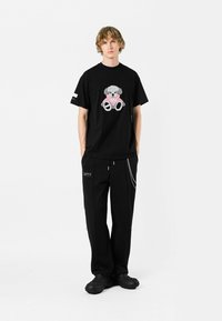 Camiseta de algodón negra con un gráfico de oso de peluche, combinada con pantalones negros que tienen un bolsillo con cremallera y detalles en cadena plateada. Zapatillas negras sin cordones.
