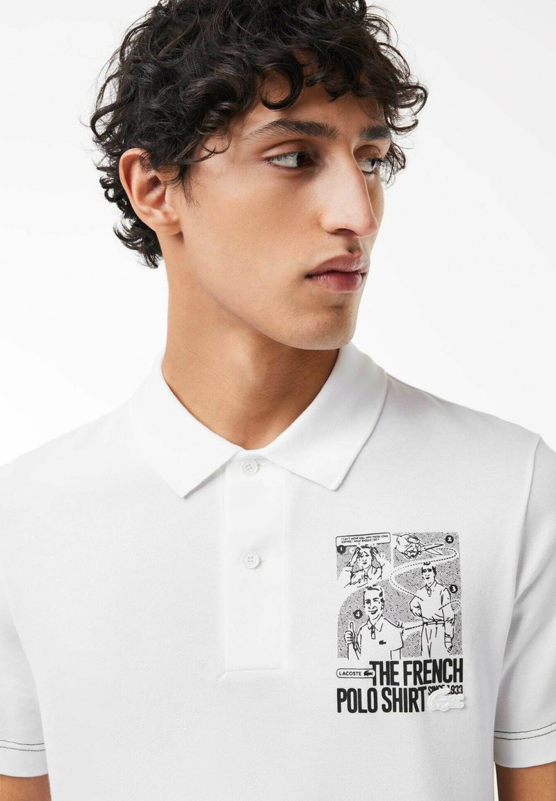 Polo blanc avec un tissu texturé. Présente un imprimé graphique noir sur la partie gauche de la poitrine avec des illustrations et du texte. Design de col classique.