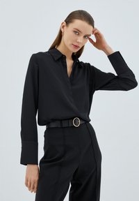 Blusa nera con colletto, maniche lunghe e dettagli bottonati, abbinata a pantaloni a vita alta e una cintura nera con una fibbia circolare dorata.