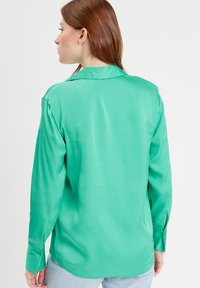 Chemise en satin vert à manches longues avec col et poignets boutonnés ; texture lisse, coupe décontractée, ourlet légèrement arrondi et détails minimaux.