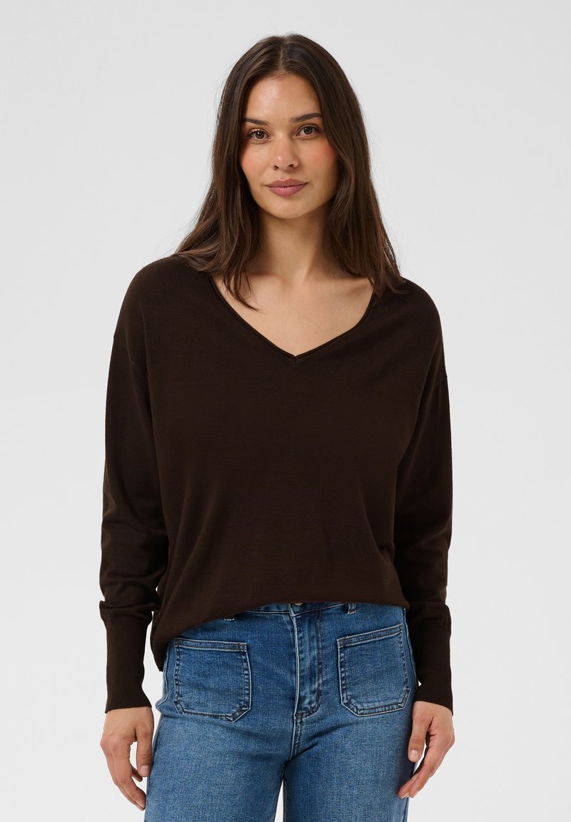 Femme aux longs cheveux foncés portant un pull marron foncé à col en V et un jean bleu, debout devant un fond clair uni.