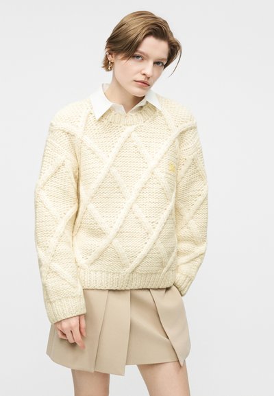 Νεαρό άτομο με κοντά μαλλιά που φοράει κρεμ πλεκτό πουλόβερ με σχέδιο κοτσίδα πάνω από λευκό πουκάμισο με γιακά και μπεζ πλισέ φούστα, στέκεται μπροστά σε απλό φόντο.