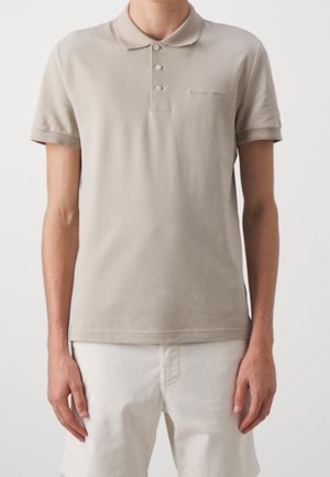 Jonge persoon die een beige poloshirt met korte mouwen draagt, los over een witte korte broek, staand tegen een effen achtergrond.