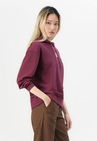 Burgundy huvtröja med en framficka, dragsko huva och en liten logotyp. Långa ärmar och avslappnad passform, tillsammans med bruna byxor.