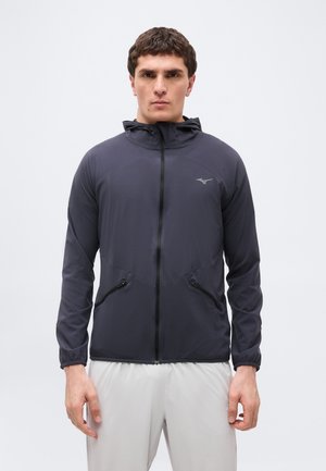 Muž v tmavě šedé bundě na zip s kapucí a zapínacími kapsami, v kombinaci se světlými šedými kalhotami, stojící před jednobarevným pozadím.