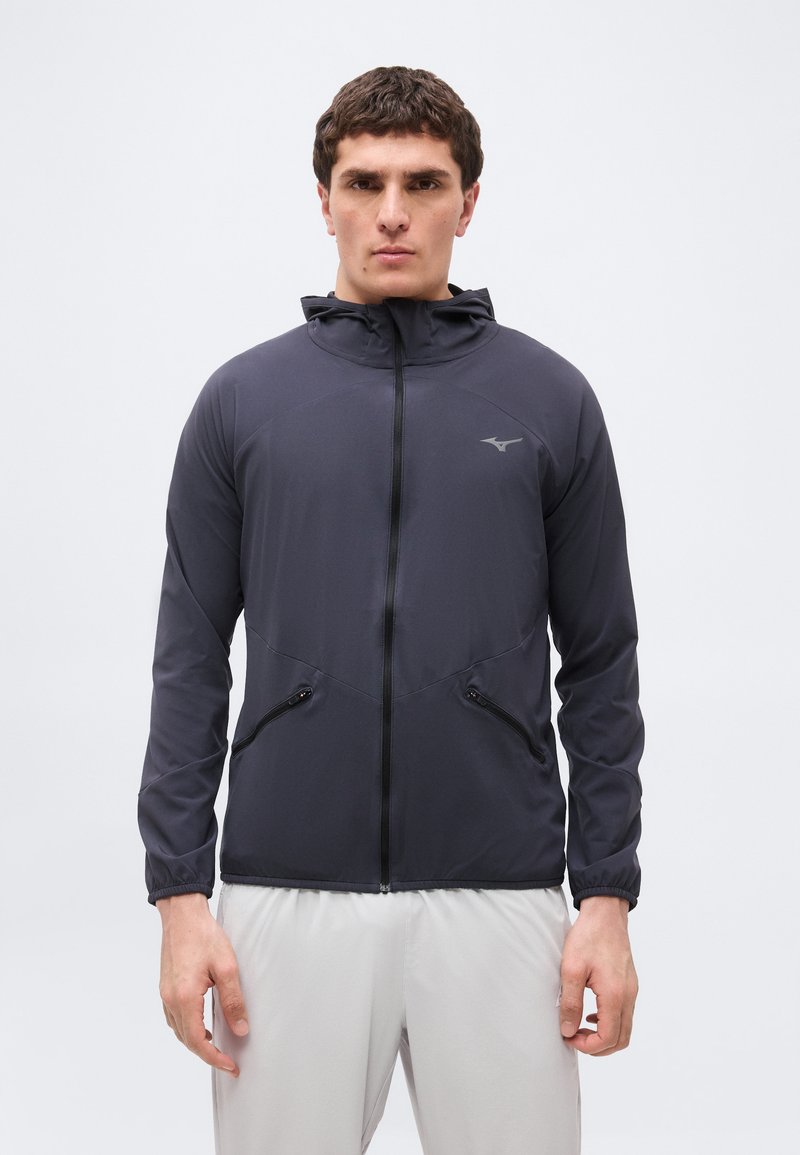 Homme portant une veste zippée gris foncé avec capuche et poches zippées, associée à un pantalon gris clair, debout devant un fond uni.