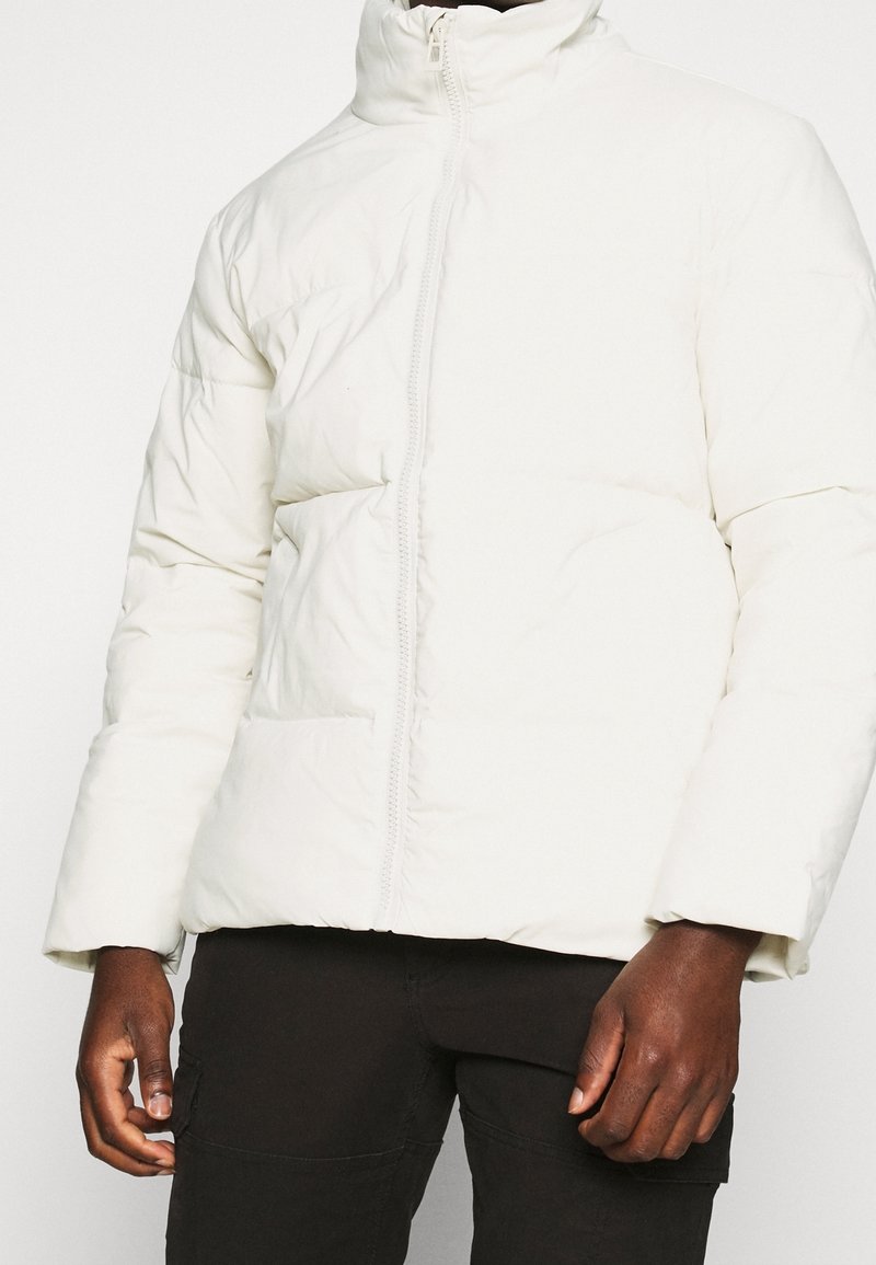 Chaqueta acolchada blanca con un cuello alto, patrón de acolchado horizontal y cremallera frontal. Presenta bolsillos laterales y un corte relajado.