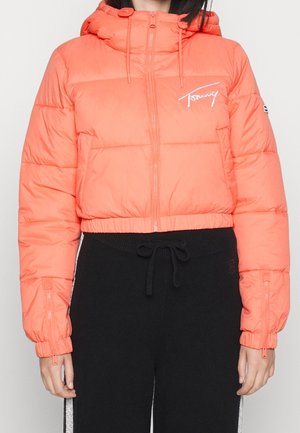 Kvinde iført en kort, lyse koral puffjakke med hætte og sorte snøre bukser med sidelister.