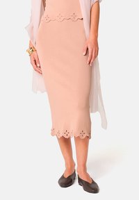 Jupe crayon beige avec ourlet festonné présentant des découpes florales. Assortie à un léger superposé blanc et des chaussures plates marron foncé.