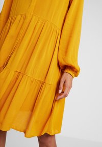 Gelbes, gewebtes Kleid mit langen Ärmeln, gerafftem Bund und gestuftem Rock, aus leichtem, strukturiertem Stoff mit Knopfdetails.