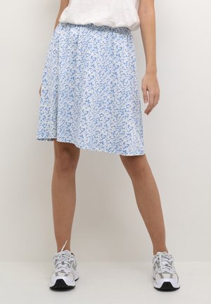 Femme portant une jupe fleurie bleue et blanche longueur genou, un haut blanc et des chaussures de sport blanches, debout devant un fond uni.