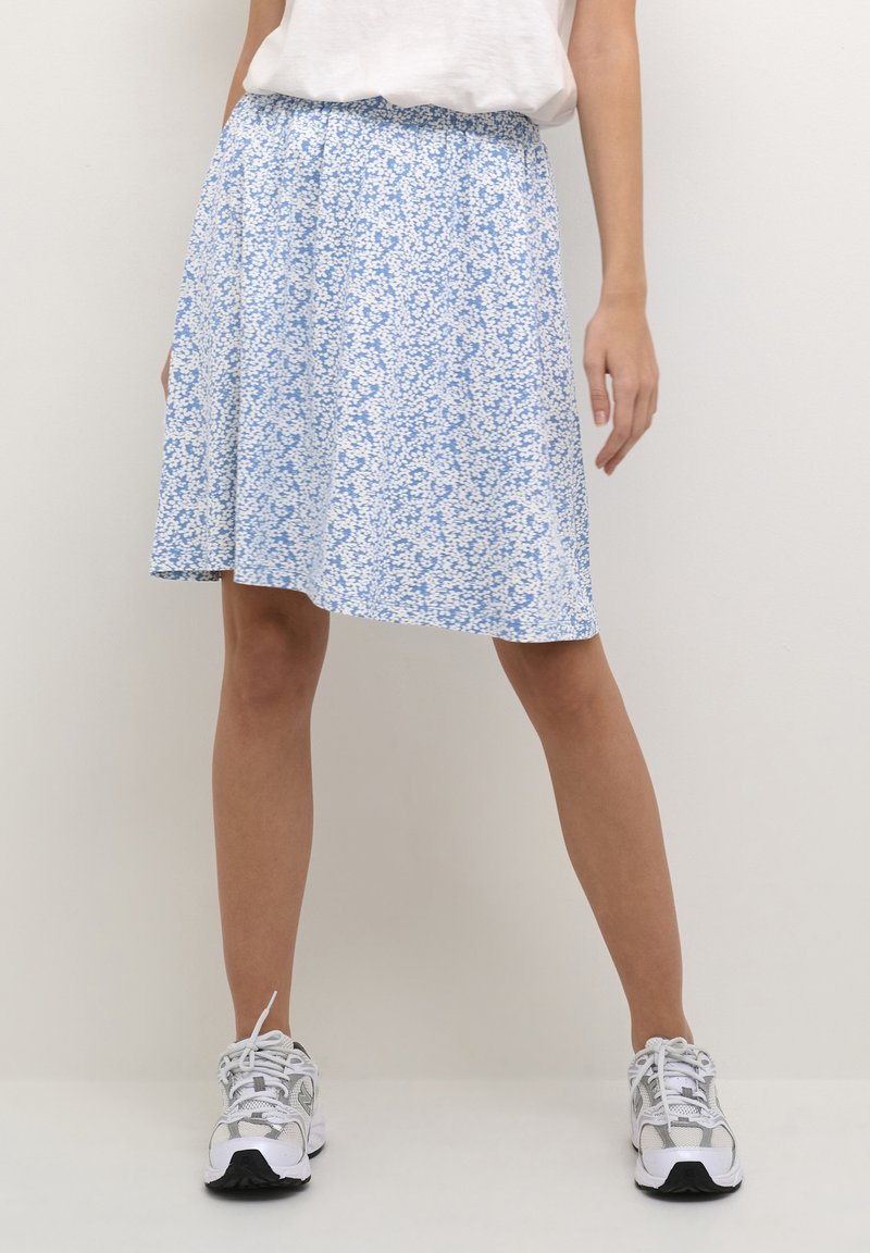 Femme portant une jupe fleurie bleue et blanche longueur genou, un haut blanc et des chaussures de sport blanches, debout devant un fond uni.