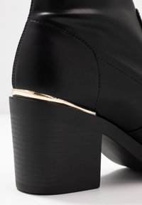 Bottine en cuir noir avec un talon bloc texturé noir et un accent métallique doré sur le talon. Fini lisse avec des détails de couture subtils.