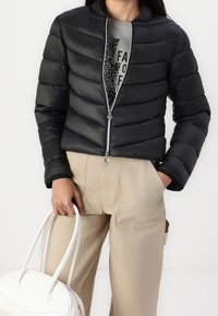 Giacca a vento nera con zip frontale, caratterizzata da un motivo a righe orizzontali e una silhouette aderente. Abbinata a pantaloni beige e una borsa bianca.